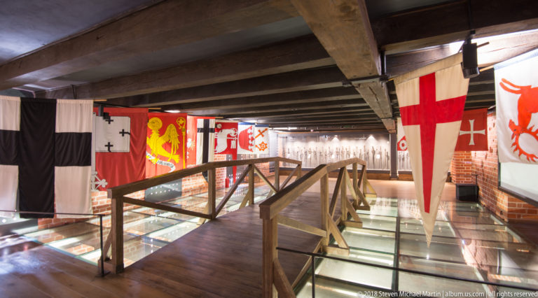 Timber gallery in a museum space hung with large heraldic banners above illuminated glass display cases.