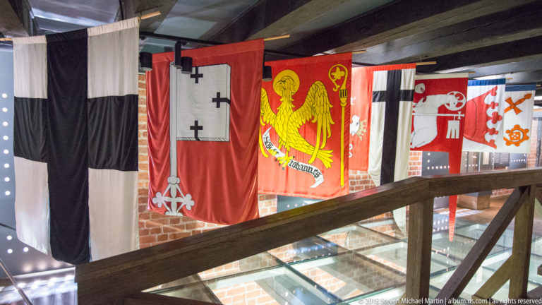 Close gallery view of large heraldic banners bearing crosses, a gold eagle, and other emblems behind a timber railing.