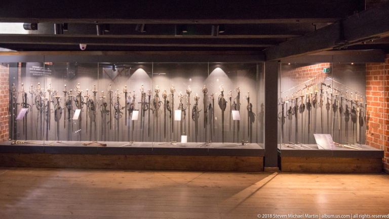 Long glass museum case displaying numerous polearms, swords, axes, and other historic edged weapons with individual labels.