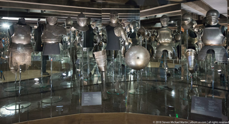 Dark museum gallery with rows of plate armor, helmets, breastplates, gauntlets, and round shields on transparent mounts.