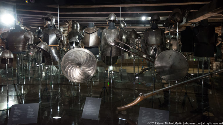 Museum display of plate-armored figures with round shields, helmets, and long polearms under dramatic spot lighting.