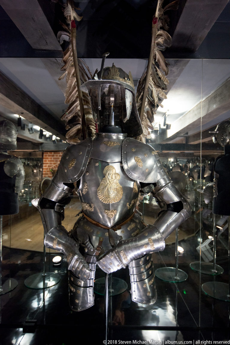 Front view of ornate dark plate armor with gilded decoration and tall feathered wings rising behind the shoulders.