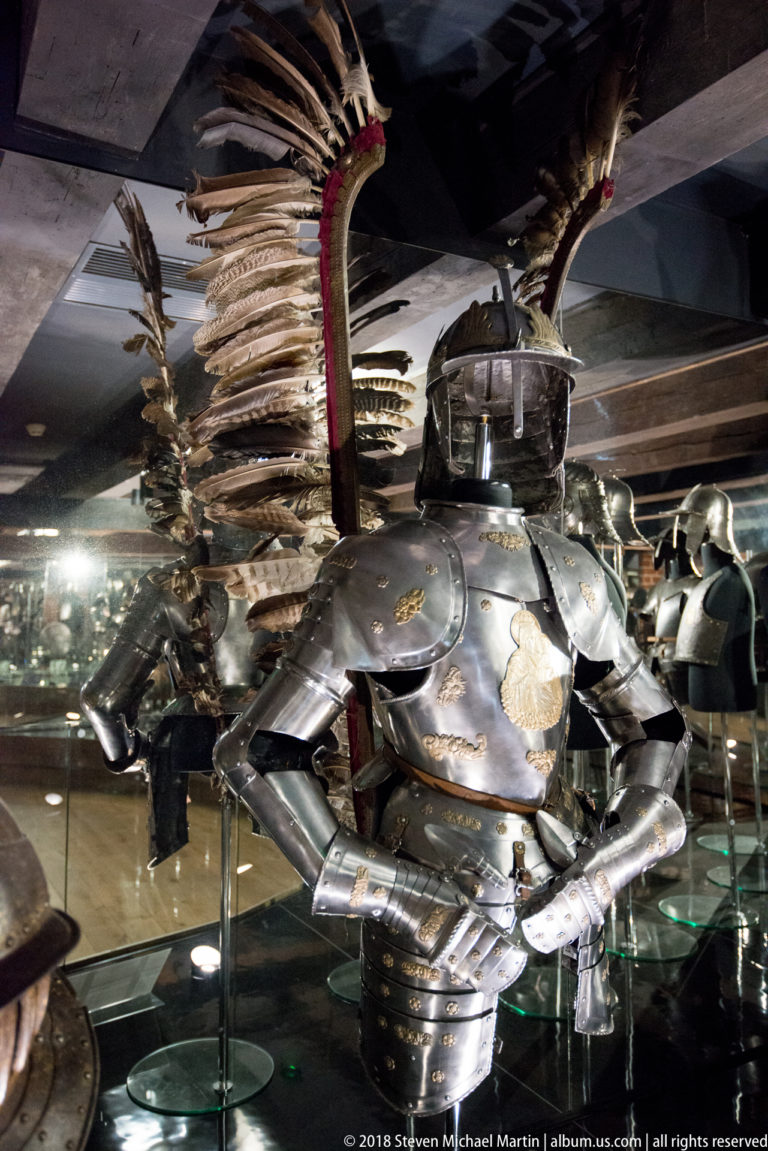 Three-quarter view of blackened plate armor with gilded ornament and large feathered wings mounted behind the shoulders.
