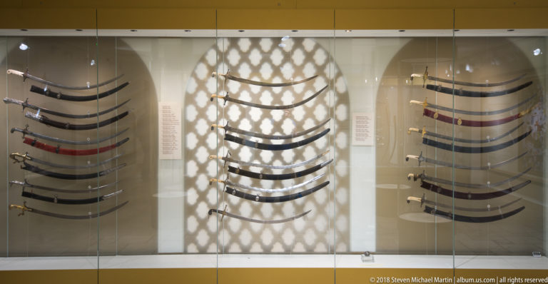 Glass museum case containing multiple curved sabres arranged in three groups, with varied hilts and scabbards.