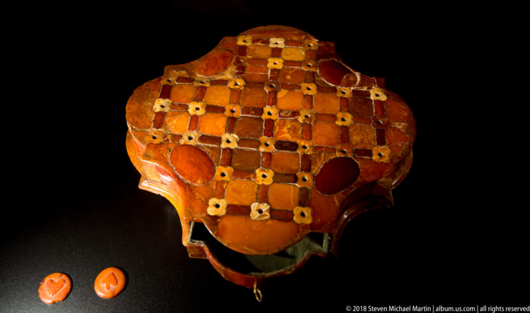 Rounded box or container covered in checkerboard amber pieces, with two loose oval amber pieces displayed beside it.
