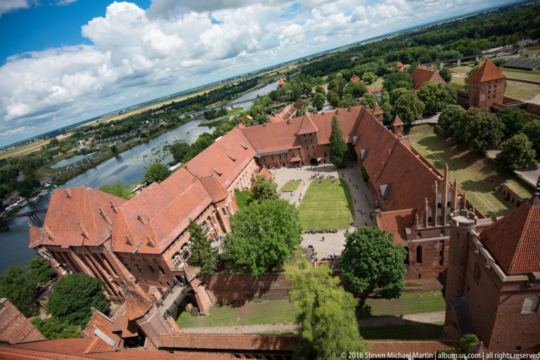High-angle view over red-tiled Malbork Castle courtyards and brick ranges toward the Nogat River and green landscape beyond.