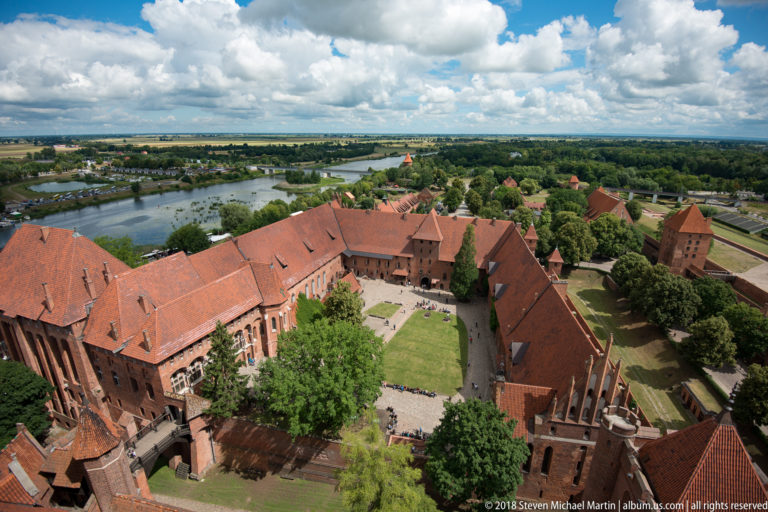 Elevated view of a broad Malbork Castle courtyard enclosed by red-brick ranges, with the Nogat River beyond.