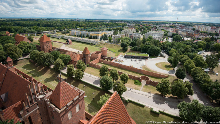 High view over Malbork Castle roofs and outer brick fortifications toward roads, trees, and the modern town beyond.
