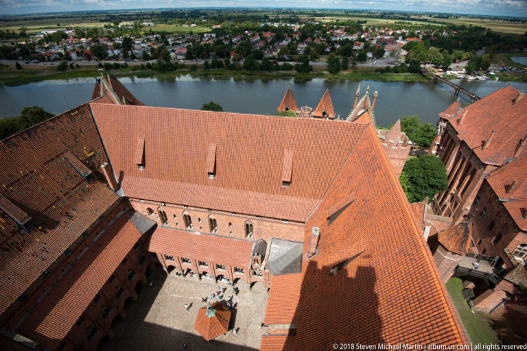 High-angle view of red-tiled castle roofs around a cobbled courtyard, with the Nogat River visible directly beyond.