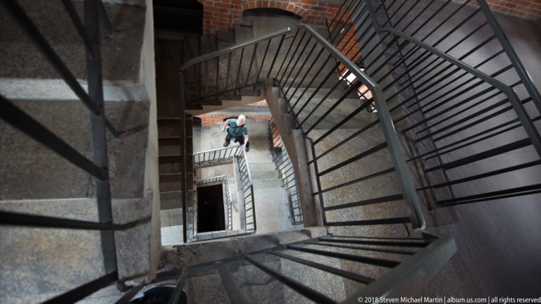 Dark metal spiral staircase descending through a stone-and-brick shaft, with a visitor visible on a lower landing.
