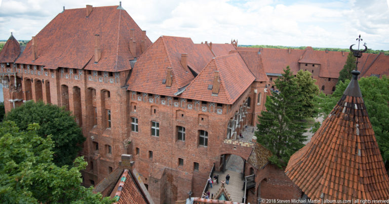 Elevated exterior view of red-brick Gothic castle ranges with steep tiled roofs, recessed arches, gables, and a conical-roofed turret.