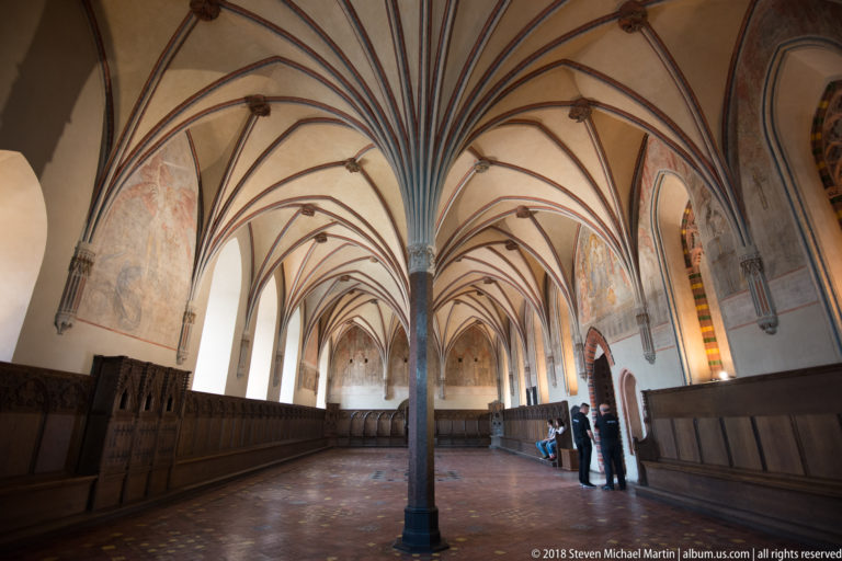 Long Gothic hall with central columns, fan-like rib vaults, faded wall paintings, dark wooden paneling, and visitors near the far end.