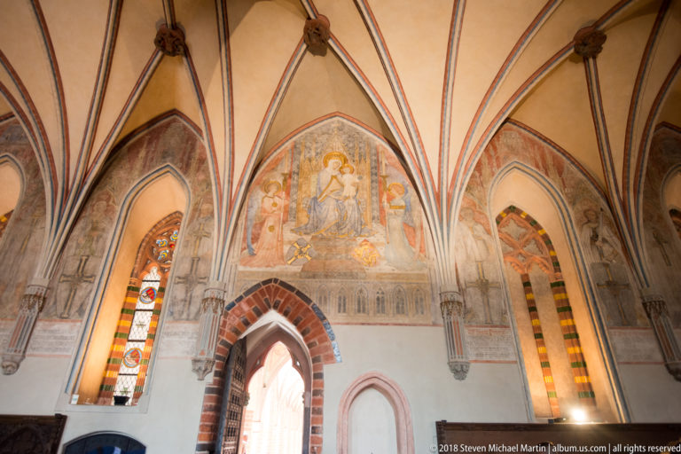 Faded religious mural above a pointed brick doorway, framed by Gothic rib vaults and narrow stained-glass windows.