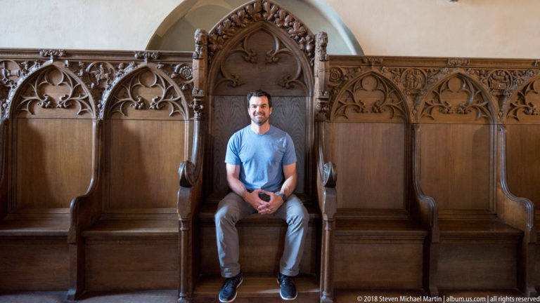 Visitor seated in a tall carved Gothic wooden stall surrounded by pointed tracery, foliage, and adjacent high-backed seats.