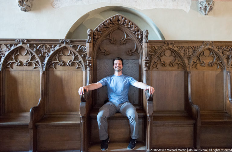 Visitor seated with arms extended on the armrests of a tall carved Gothic wooden stall with decorative tracery.