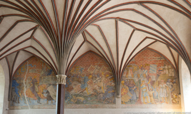 Large faded narrative murals of mounted and standing figures beneath radiating Gothic rib vaults and a central column.