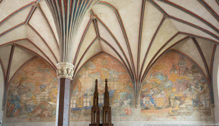 Dark pinnacled Gothic wooden furnishing standing before large narrative wall murals under radiating vault ribs.