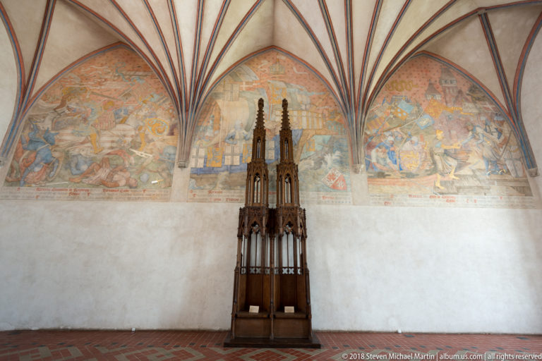 Tall dark Gothic wooden furnishing with pinnacles and tracery standing beneath two large faded narrative wall paintings.
