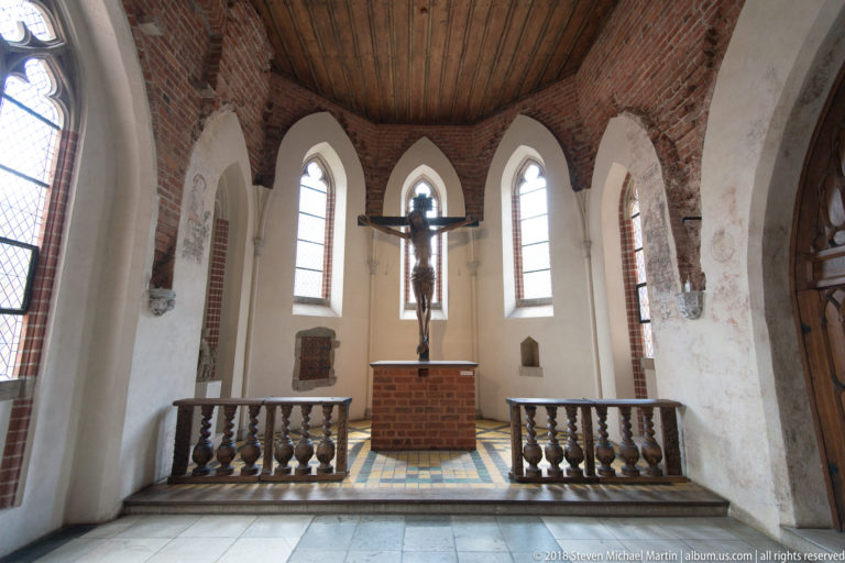 Small chapel with a brick altar, hanging crucifix, pointed windows, white plaster walls, and low wooden sanctuary rail.