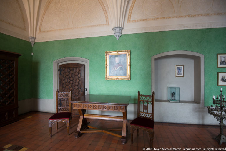 Green-walled historic room with a wooden bench, two carved chairs, framed portraits, and a pale decorated vaulted ceiling.