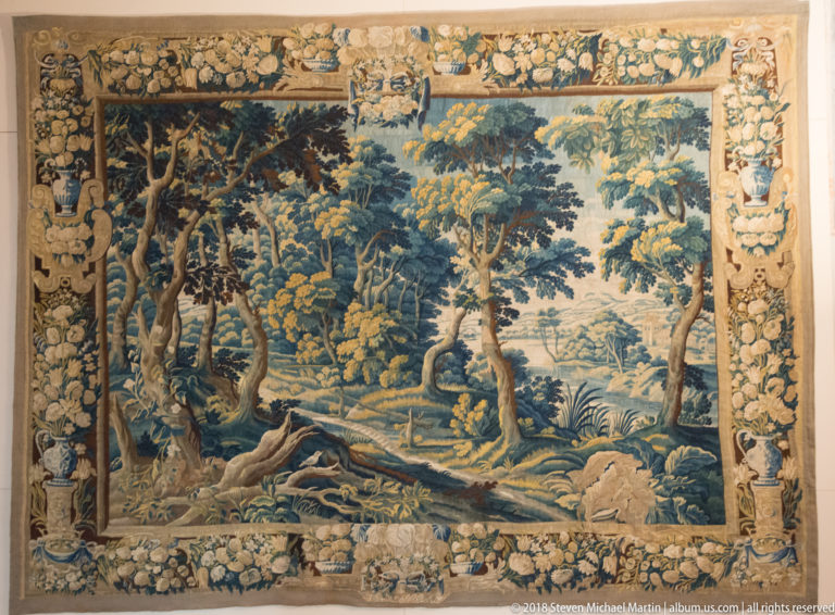 Large tapestry of a wooded landscape with stream, animals, dense trees, and an ornate floral border.