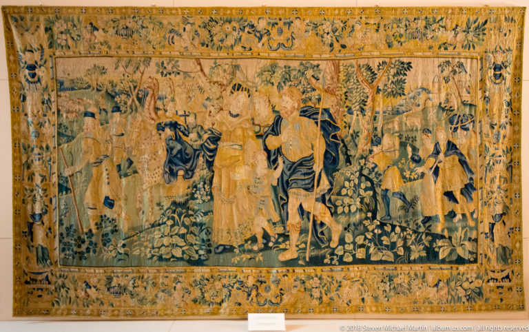 Large tapestry showing richly dressed courtly figures among flowers and foliage within a broad ornamental border.