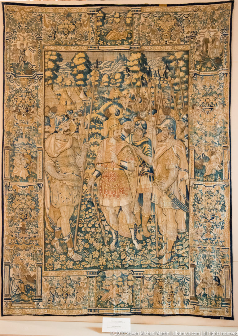 Large woven battle tapestry showing mounted soldiers with long spears moving through a wooded landscape.