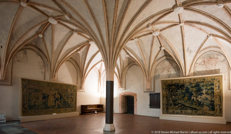 Vaulted Gothic chamber with central column and two large tapestries, one figural and one a wooded landscape.