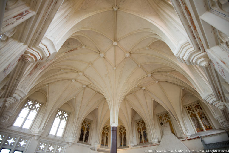 Upward view of an ornate pale Gothic rib vault with carved bosses, traceried windows, and narrow painted wall niches.