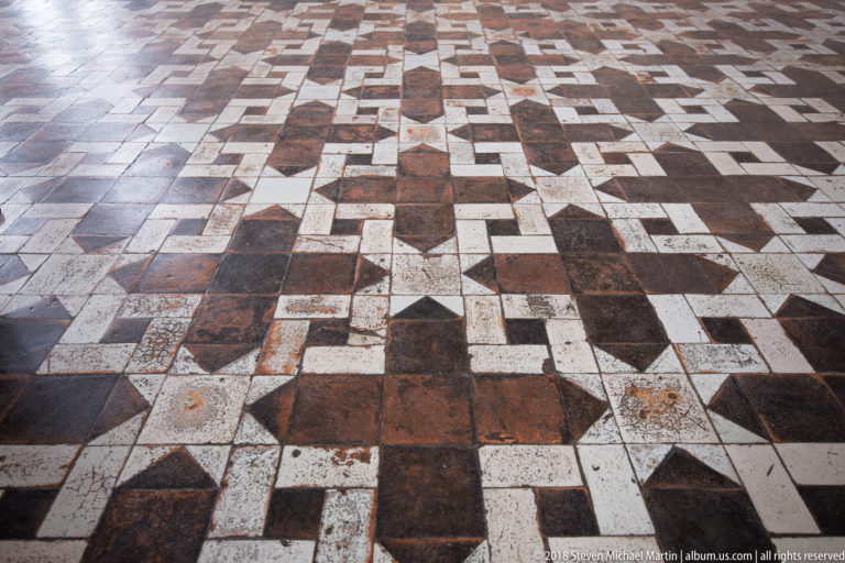Close view of worn pale and dark rectangular floor tiles arranged in interlocking geometric cross and stepped patterns.
