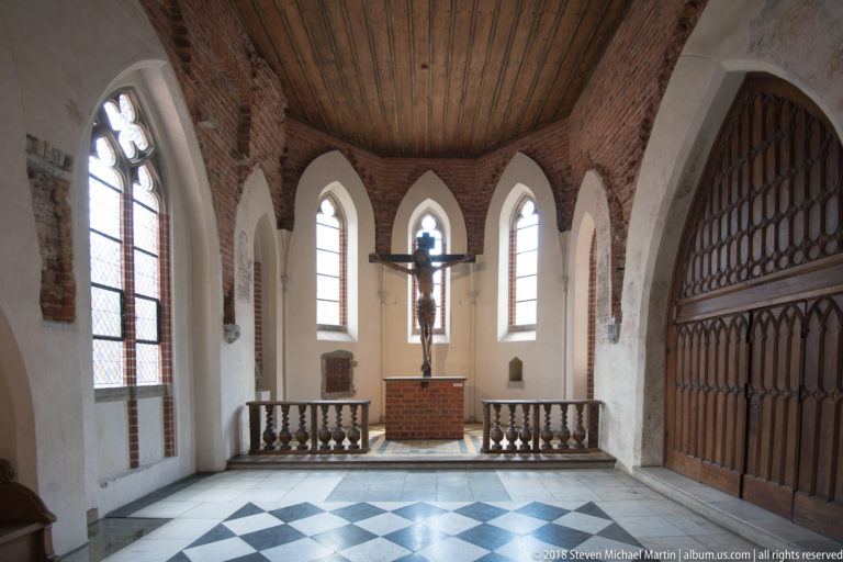 Small chapel with crucifix and brick altar beyond a black-and-white checkered floor, pointed windows, and a large wooden screen.