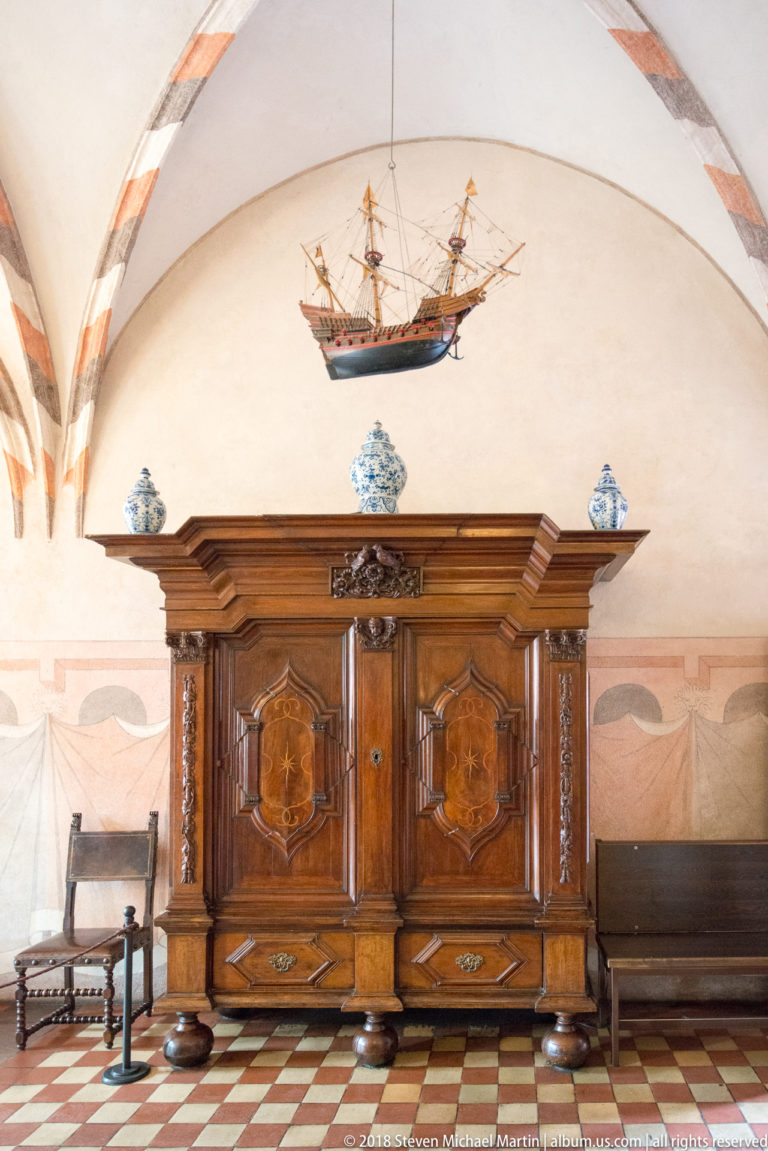 Ornate historic wooden cabinet with blue-and-white ceramics beneath a suspended model sailing ship in a furnished room.
