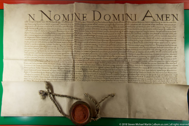 Large parchment charter headed “IN NOMINE DOMINI AMEN” with dense handwriting and a red wax seal suspended on cords.
