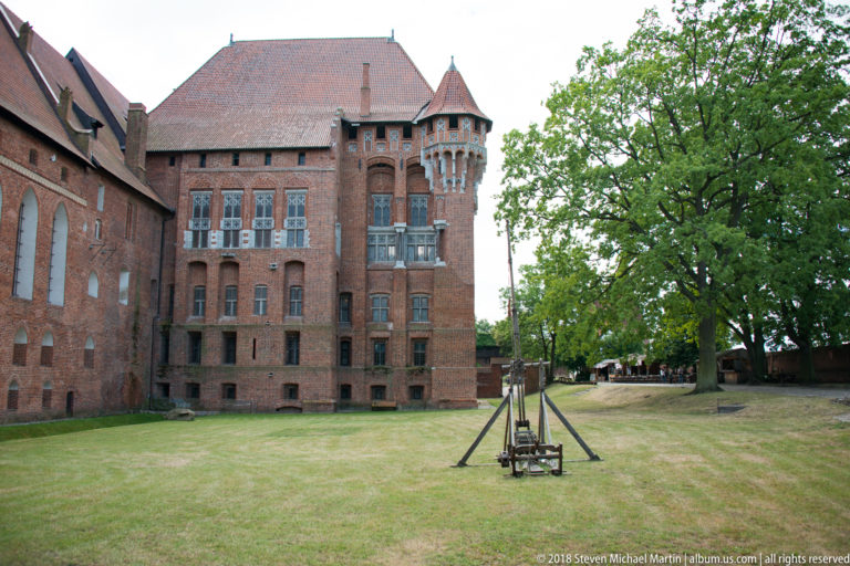 Red-brick castle range with projecting tower beside a broad grassy courtyard, mature tree, and small timber structure.