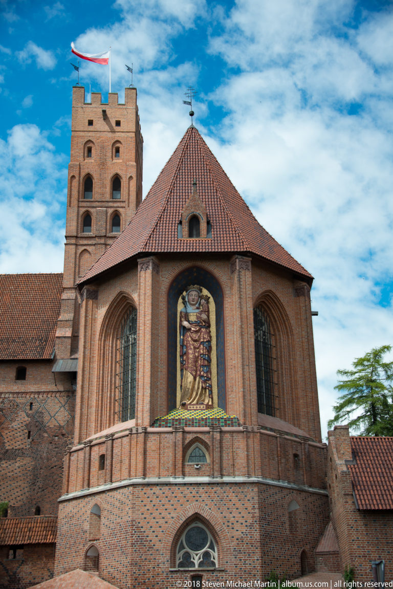 Brick Gothic church façade with tall pointed windows and a monumental Virgin and Child mosaic beneath a steep tiled roof.