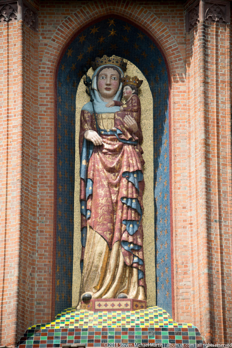 Full-length mosaic of the crowned Virgin holding the Christ Child in a star-filled blue arched niche framed by red brick.