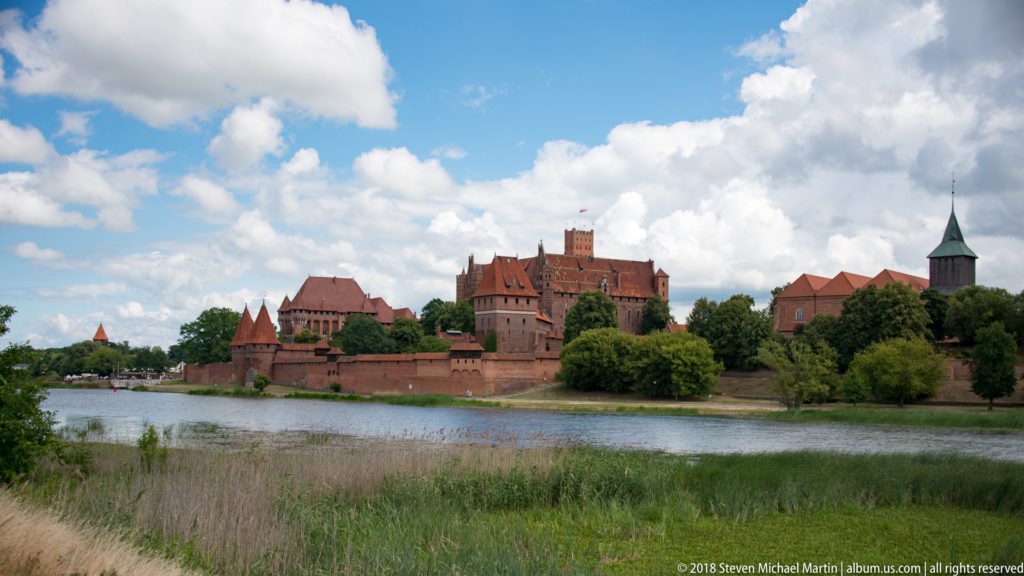 Wide view of Malbork Castle across the Nogat River, with red-brick towers and roofs beyond reeds, trees, and a riverside path.