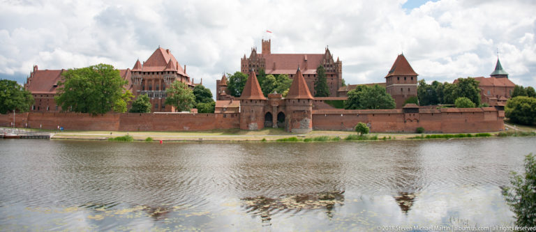 Frontal view of Malbork Castle across the Nogat River, with fortified walls, gate towers, higher brick ranges, and water reflections.