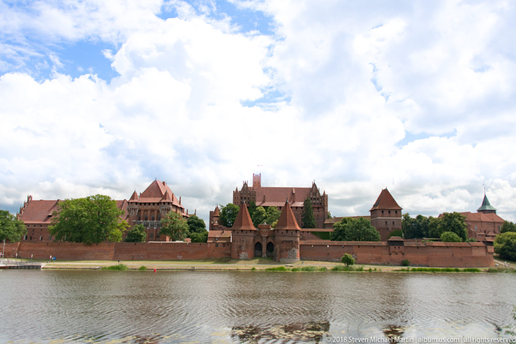 Broad panorama of Malbork Castle across the Nogat River with long brick walls, towers, upper ranges, and a cloud-filled sky.
