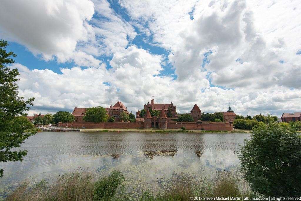 Distant panoramic view of Malbork Castle across the Nogat River, framed by reeds and trees beneath large cumulus clouds.