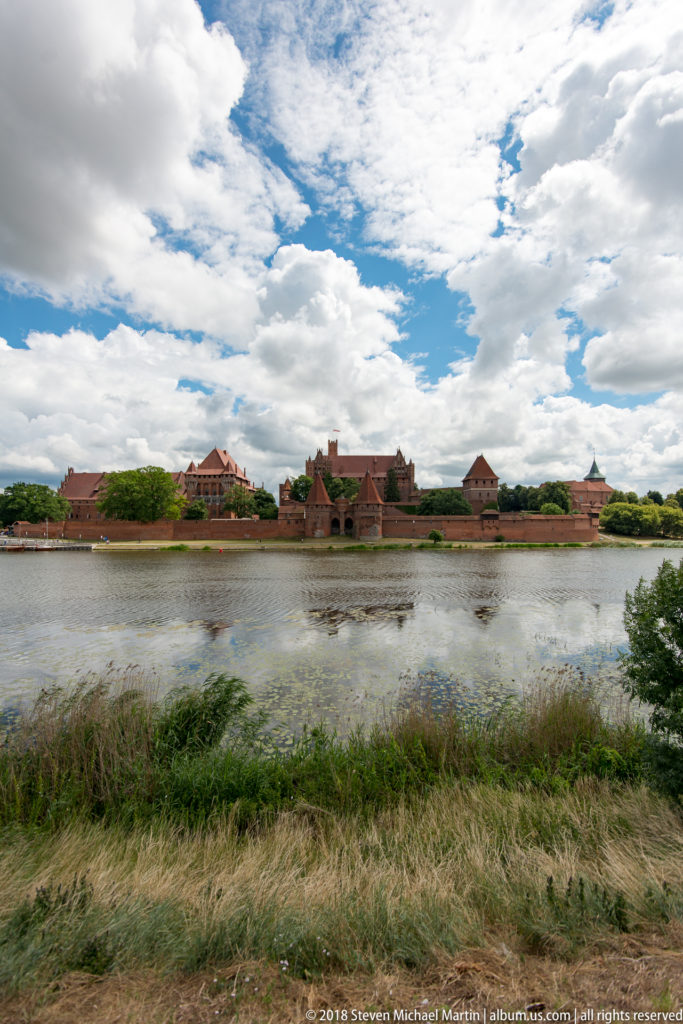 Very wide view of Malbork Castle across the Nogat River, with foreground grasses and an expansive sky filled with clouds.