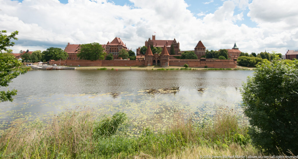 Malbork Castle across the Nogat River, with brick walls and towers framed by trees and aquatic plants in the foreground.