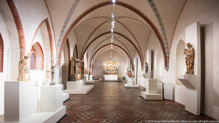 Long vaulted gallery with medieval religious sculptures on white plinths and an illuminated altarpiece at the far end.