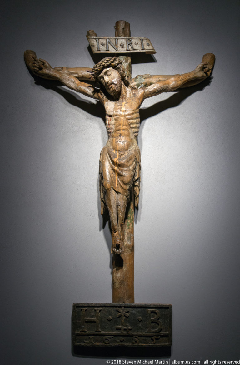 Weathered polychrome wooden crucifix with Christ on a dark cross beneath an INRI tablet, displayed against a plain wall.