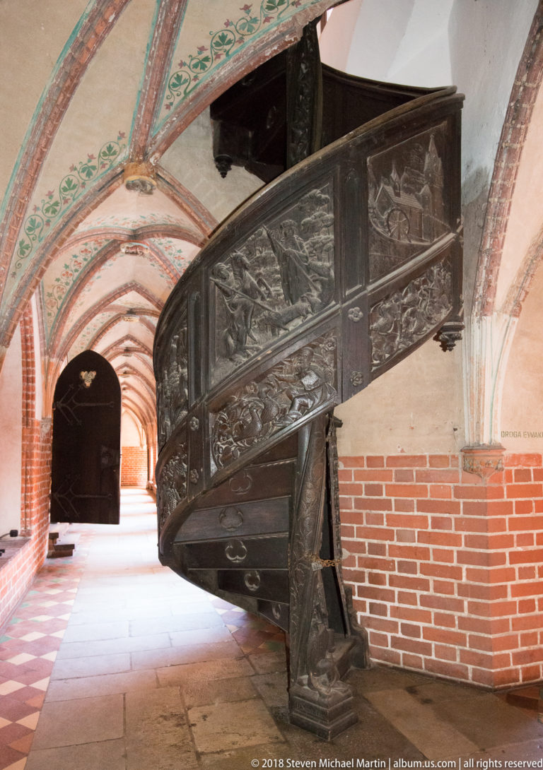 Curving dark wooden staircase with carved narrative relief panels in its balustrade, set within a brick-vaulted passage.