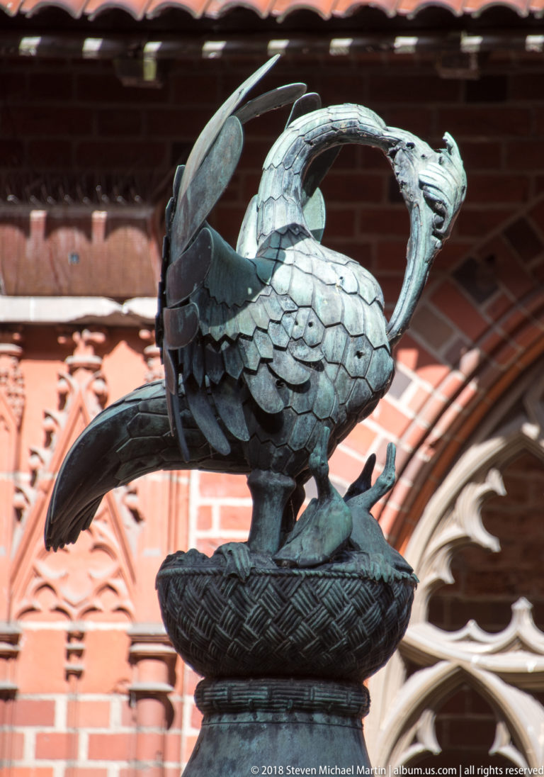 Patinated metal dragon with raised wings, long tail, and open jaws perched on a circular base before red brick and Gothic stonework.