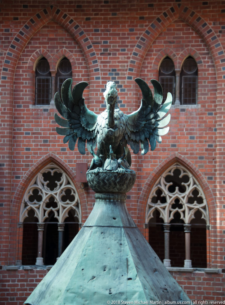 Patinated metal bird with wings fully spread atop a sloping roof, framed by Gothic tracery windows in a red-brick wall.