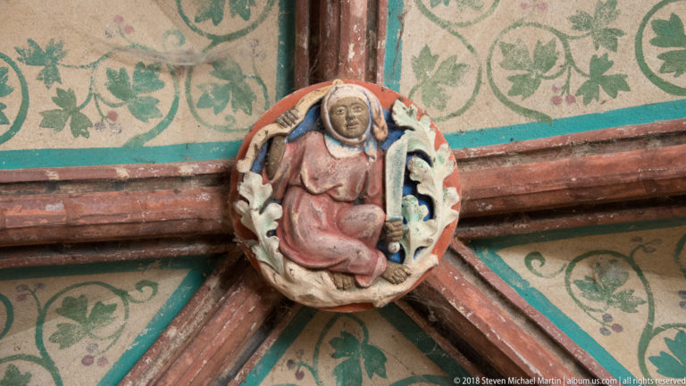 Small painted female figure set at the crossing of vault ribs, surrounded by green foliate decoration on pale plaster.