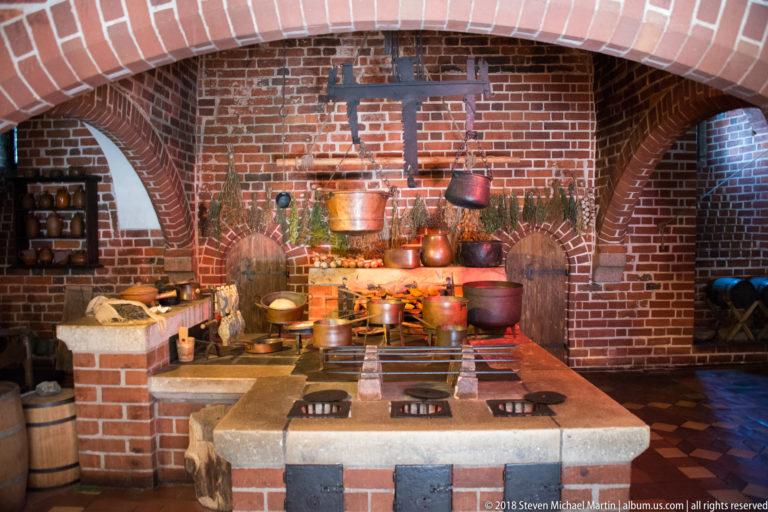 Historic brick kitchen with a broad cooking range, metal grates, hanging copper pots, kettles, shelves, and arched recesses.