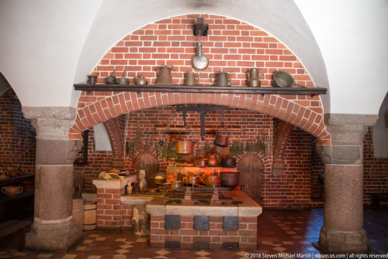 Wide historic kitchen view with brick cooking range, copper vessels, shelves, large masonry arch, stone columns, and patterned tile floor.